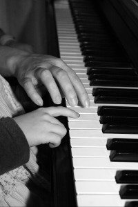 Handen van een ouder en kind samen achter het toetsenbord van een piano. 